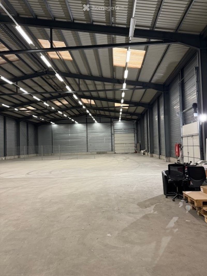 Spacious warehouse with a high ceiling, industrial lighting, a large open area, a white door, and some office chairs and pallets in the corner.