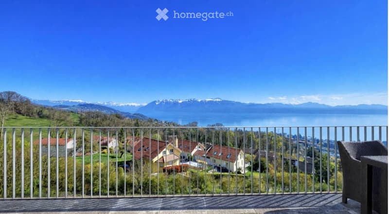 Balcony with a metal railing overlooking a scenic landscape of houses, trees, and a distant lake and mountains.