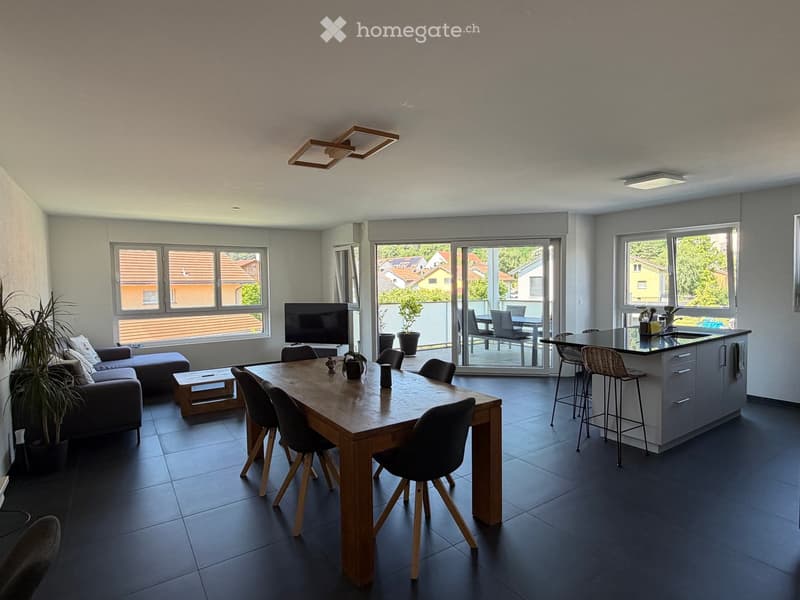 Open-plan living and dining area with a sofa, coffee table, dining table, chairs, kitchen island, and television. Large windows and sliding glass doors lead to a balcony with outdoor furniture.