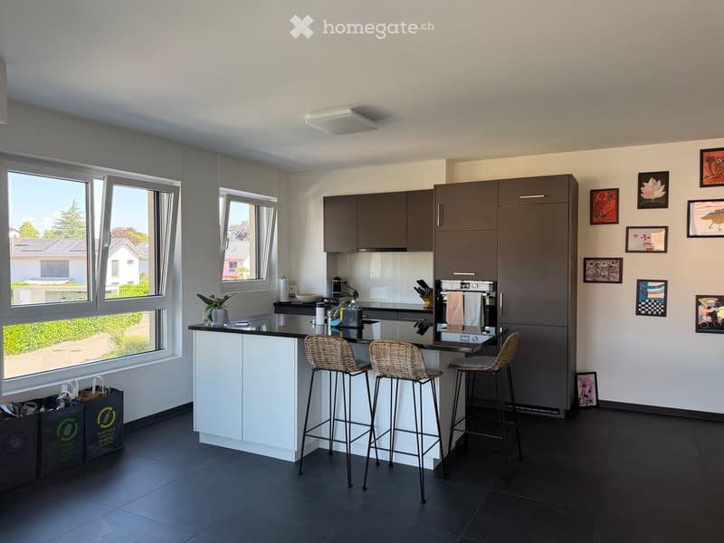 Modern kitchen with grey cabinets, white island, black countertop, three bar stools, and various appliances. Large windows provide natural light, and framed pictures decorate the wall.