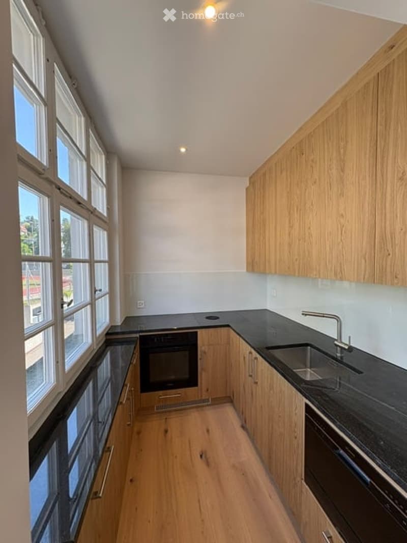 Modern kitchen with wooden cabinets, black countertops, and stainless steel appliances. Large windows provide natural light.