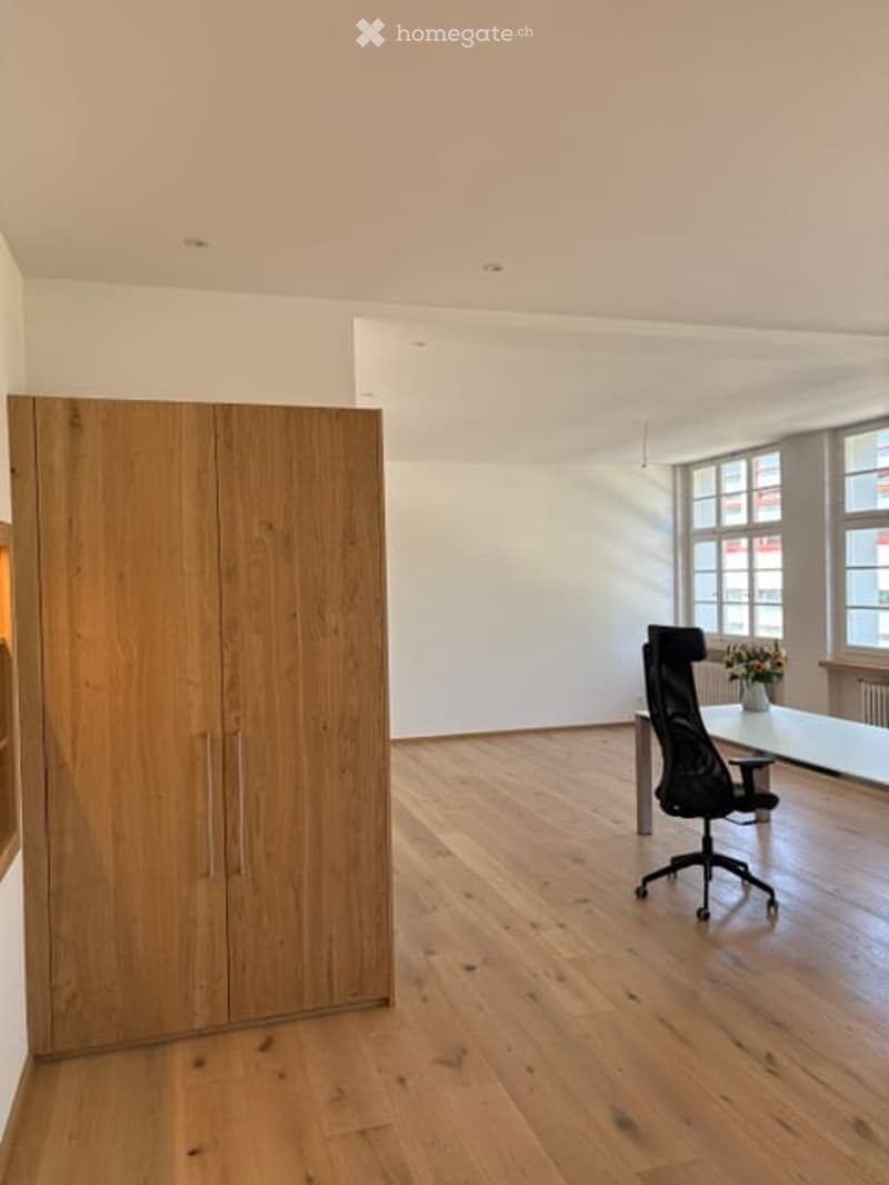 Bright, minimalist room with wooden flooring, large windows, a tall wooden wardrobe, a black office chair, and a small table with a vase of flowers.
