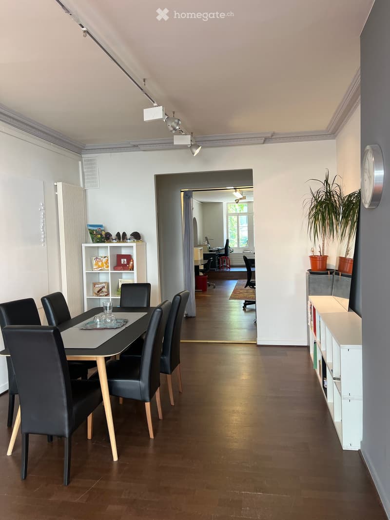 Dining area with a black dining table and six black chairs, a white shelf with books and decorative items, a potted plant, and a view into an adjacent office space with a desk and office chair.