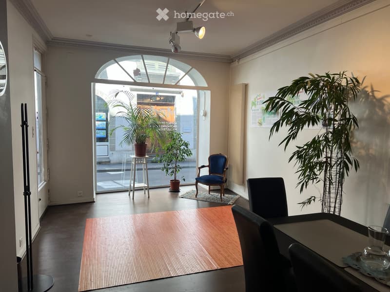 Brightly lit room with a large arched window, a wooden table, chairs, a small stool, and potted plants.