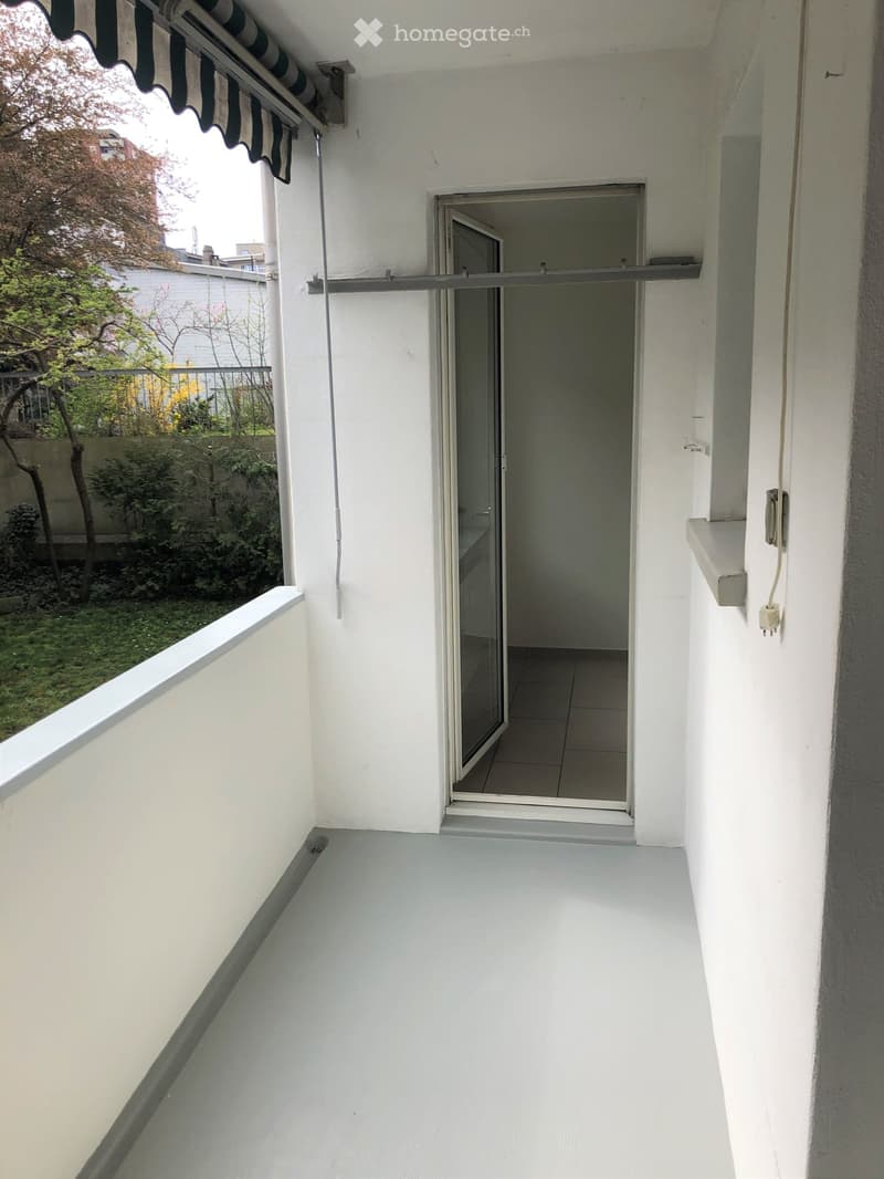 Balcony with white walls, gray floor, retractable awning, glass door leading to another room