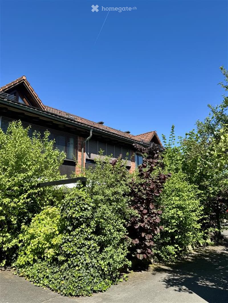 Building. A two-story residential building with a red-tiled roof, surrounded by lush green trees and shrubs. The building features multiple windows and a balcony on the upper floor.
