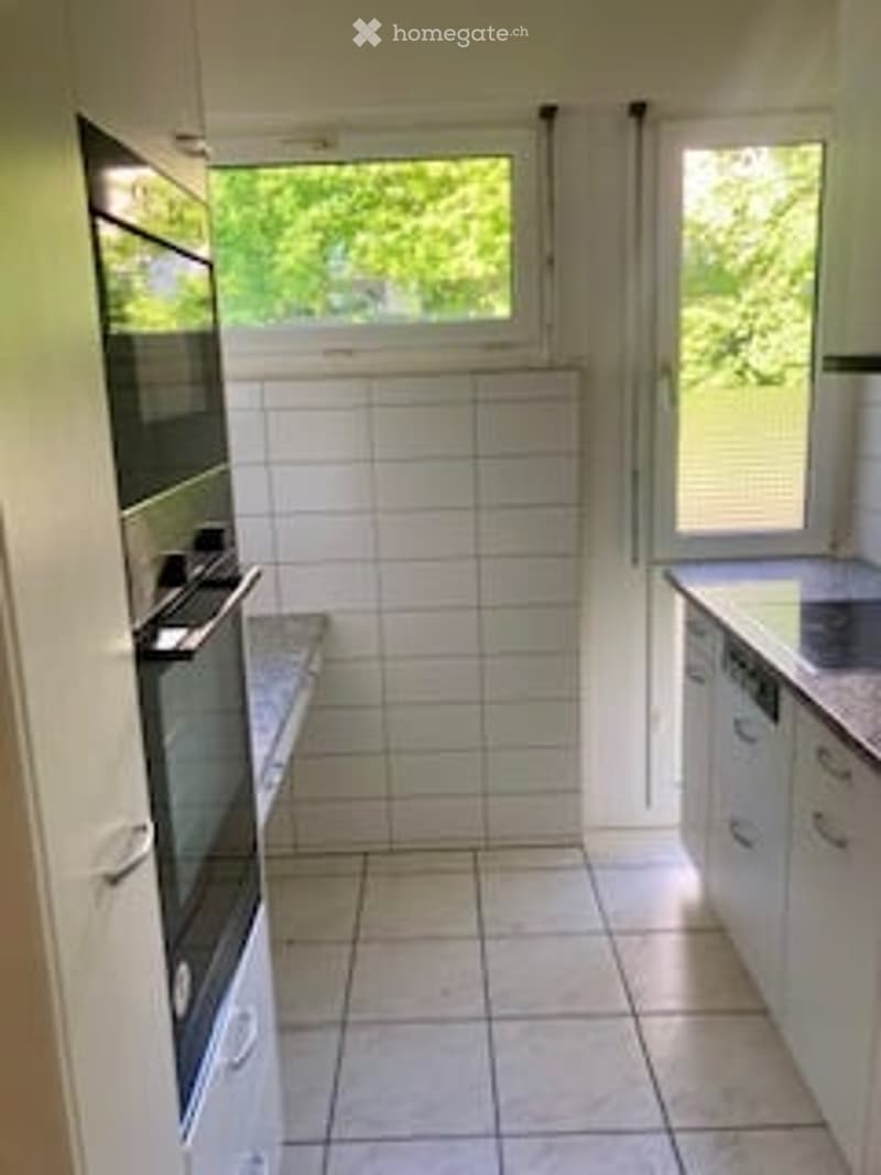 Modern kitchen with white tiled walls, white cabinets, a built-in oven, and a dishwasher. The space is clean and well-lit with natural light from the windows.