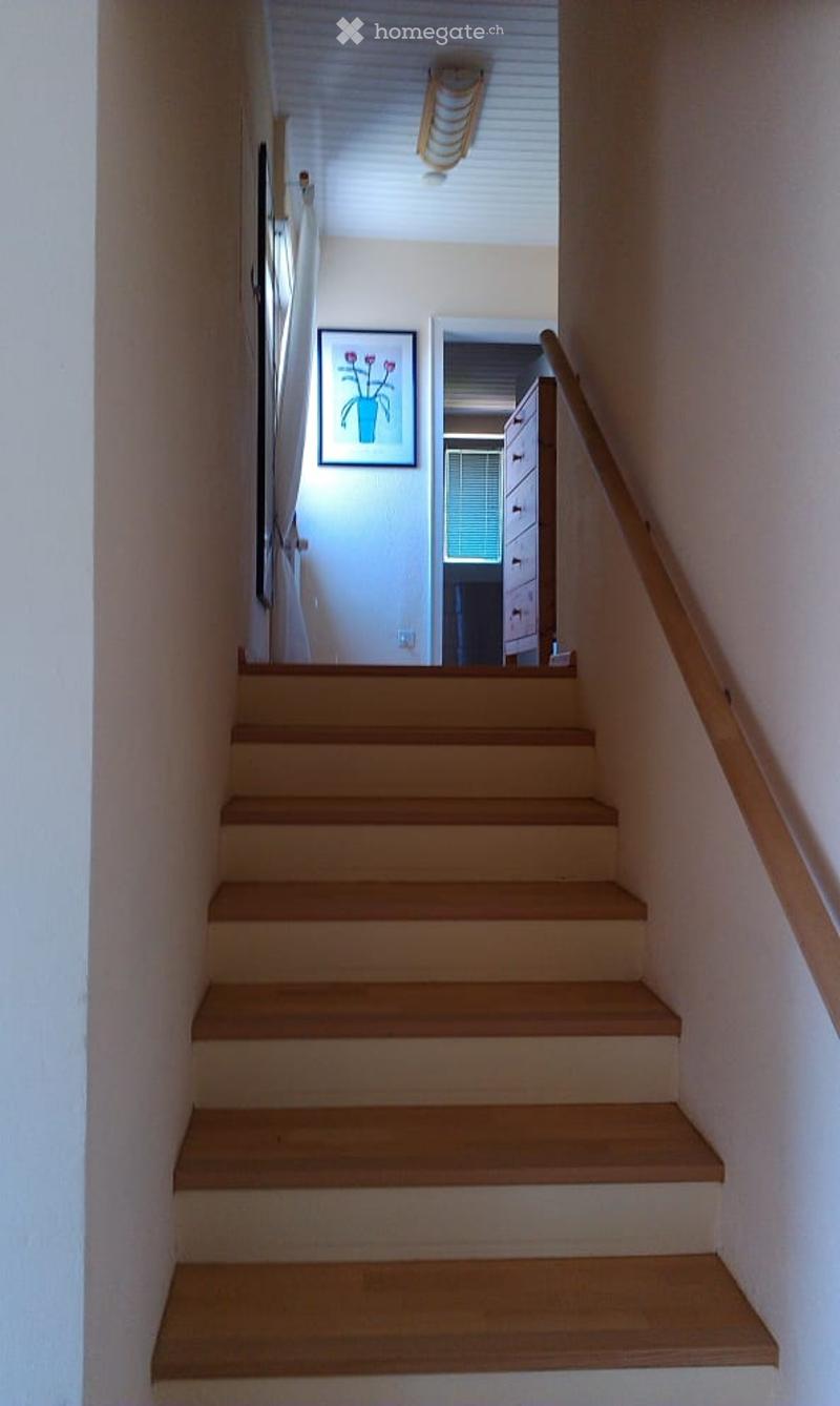 Wooden staircase with a handrail, leading to an upper level. The walls are painted in a light color, and there is a framed picture on one wall.