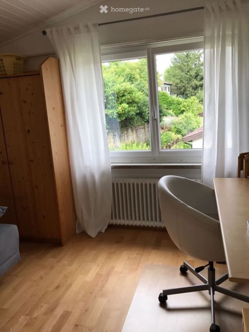 Bright room with wooden flooring, large window with white curtains, radiator below, and a simple chair and table setup.
