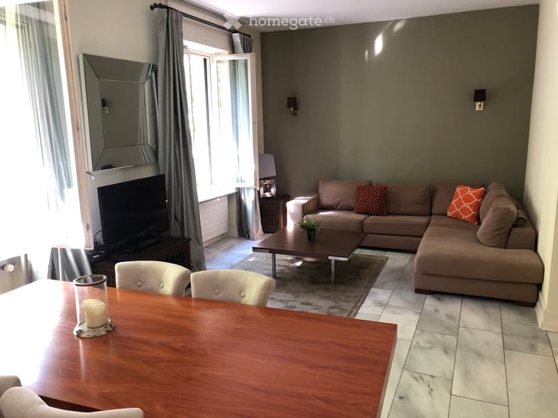 Modern living room with a sectional sofa, coffee table, dining table, chairs, and a TV. Features large windows and a balcony door.