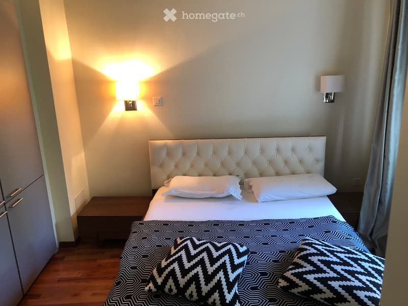 Modern bedroom with a tufted headboard bed, white bedding, black and white geometric throw pillows, and a wooden nightstand with a wall-mounted lamp.