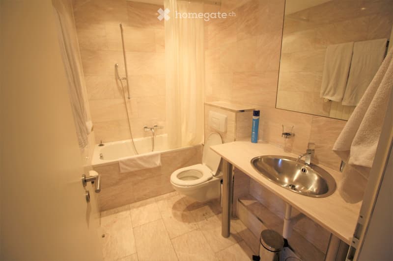 Modern bathroom with a bathtub, toilet, sink, and mirror. Light-colored tiles and a towel hanging on the side.