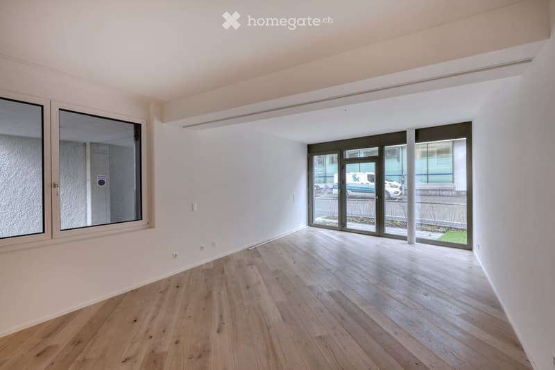 Empty room with light wooden flooring, white walls, large windows, and a set of glass doors leading outside.
