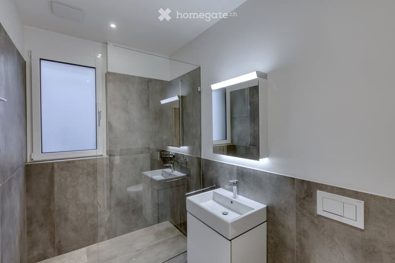 Modern bathroom with two sinks, a large mirror with built-in lighting, a glass-enclosed shower, and a toilet. The walls are tiled in a neutral gray color, and the floor is made of light-colored tiles.