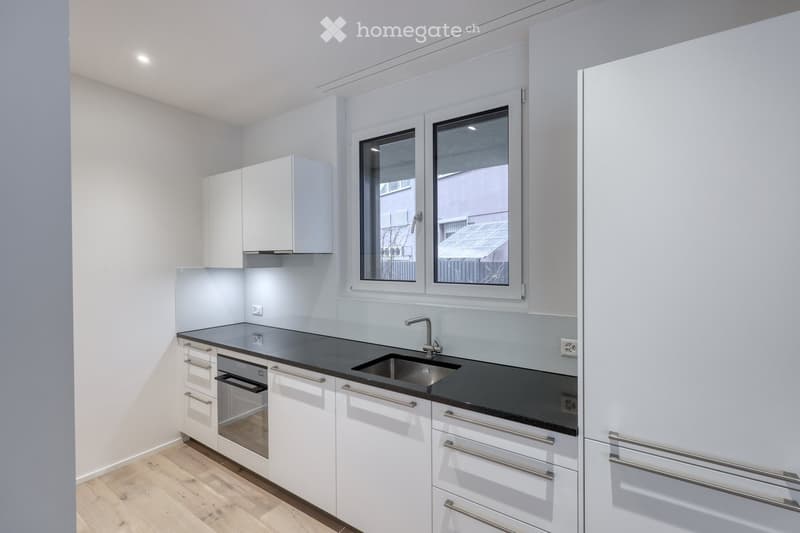 Modern kitchen with white cabinets, black countertops, and a stainless steel sink. Features include a built-in oven, ample storage, and large windows providing natural light.