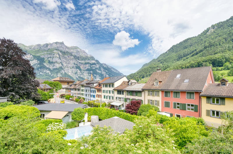 A picturesque village nestled in a valley with traditional houses, surrounded by lush greenery and towering mountains under a partly cloudy sky.
