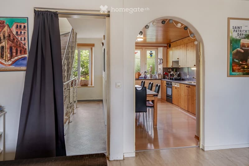 A modern hallway with a dark curtain on the left, leading to a bright kitchen area with wooden cabinets, a dining table, and chairs. The hallway features a sleek metal railing and a large arched opening.
