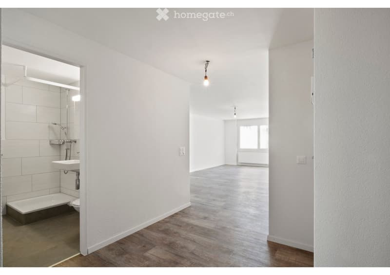 Empty room with wooden flooring and white walls, featuring a small bathroom area with a sink, bathtub, and toilet.