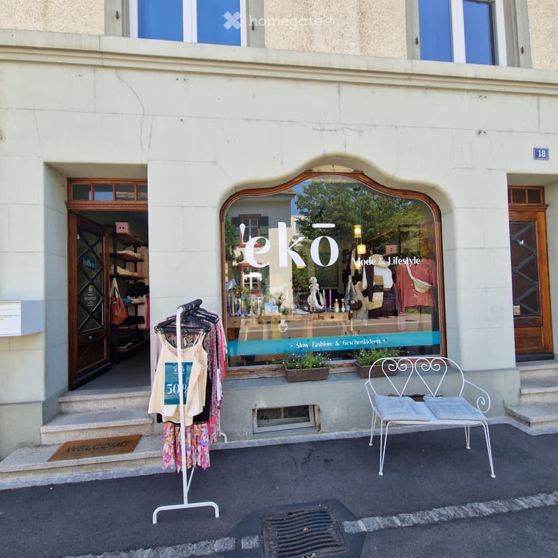 Facade of a two-story building with a storefront labeled "eko" featuring fashion and lifestyle items. Visible elements include a clothing rack with garments, a white metal chair, and a "WELCOME" doormat.