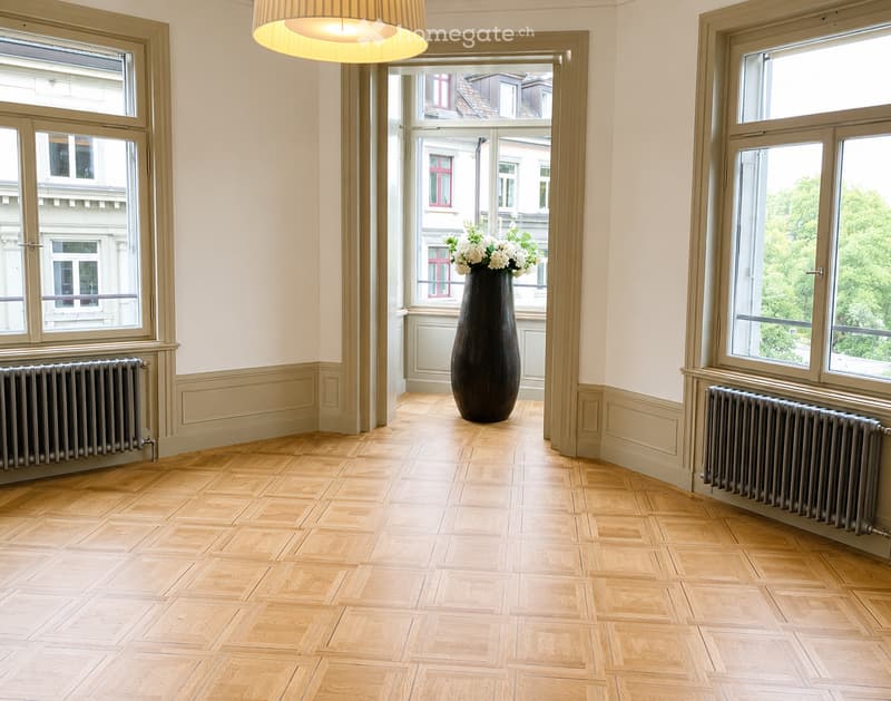 Empty room with wooden herringbone flooring, large windows, radiators, and a tall vase with white flowers in the center.
