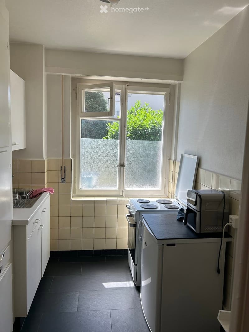 Compact kitchen with white cabinets, black countertop, and tiled backsplash. Features an oven, microwave, and refrigerator, with a window offering a view of greenery.