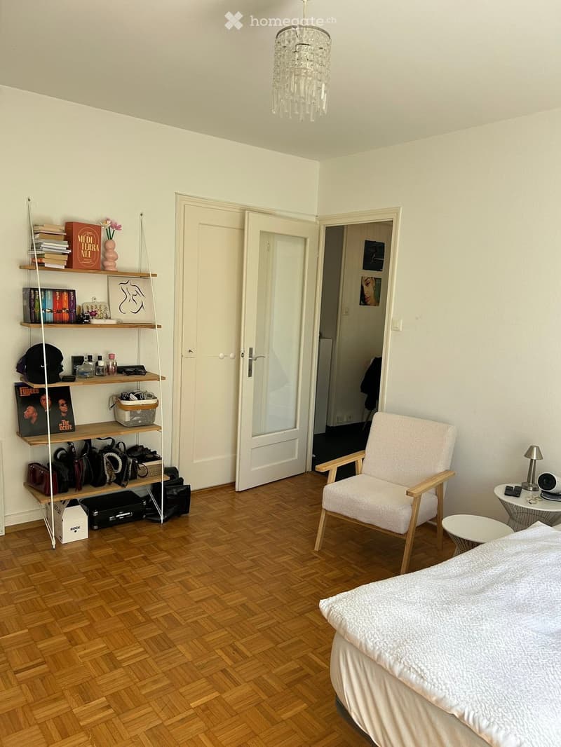 Cozy bedroom with wooden flooring, a bed with white bedding, a white armchair, a small round table with a lamp, and a tall shelf holding books, bags, and decorative items.
