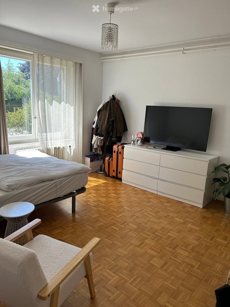 Modern bedroom with wooden herringbone flooring, large window with sheer curtains, bed with grey duvet, white side table, armchair, TV on dresser, coat rack with jackets, and potted plant.