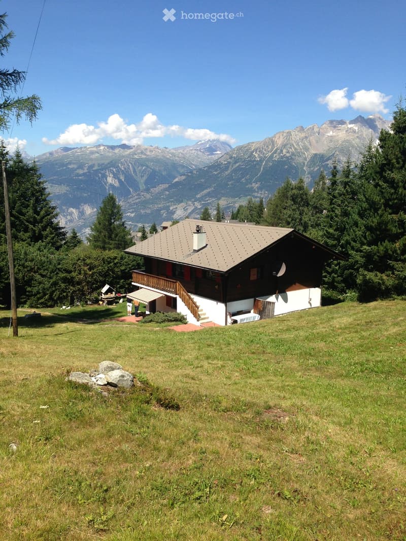 A two-story wooden house with a gray roof and white walls, situated on a grassy slope with a view of mountains in the background.