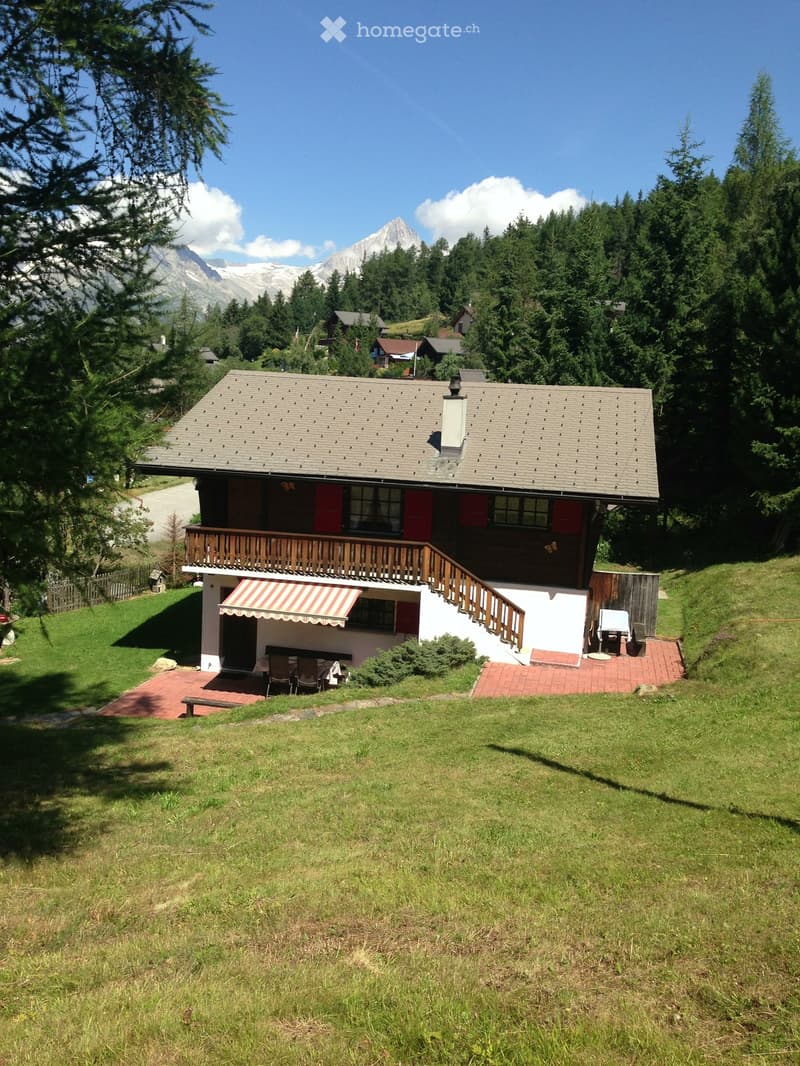 A two-story wooden house with a gray tiled roof, red shutters, and a balcony. The house is surrounded by a well-maintained lawn and features outdoor seating areas with tables and chairs.