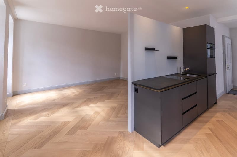 Modern kitchen with dark grey cabinets, a black countertop, and a double sink. The floor is made of light wooden parquet, and the walls are painted in a light grey color. The kitchen features built-in storage and a sleek, contemporary design.