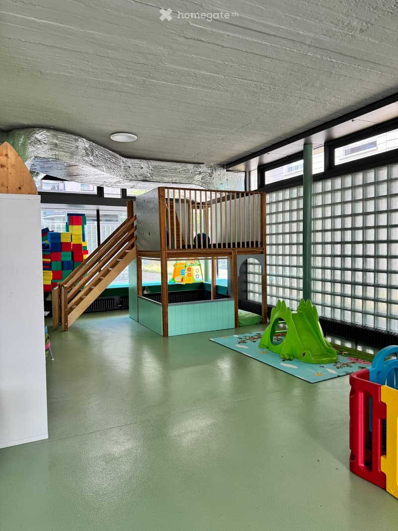 Bright, open play area with green flooring, staircase leading to an elevated play structure, colorful toy blocks, play mat, and a green slide.
