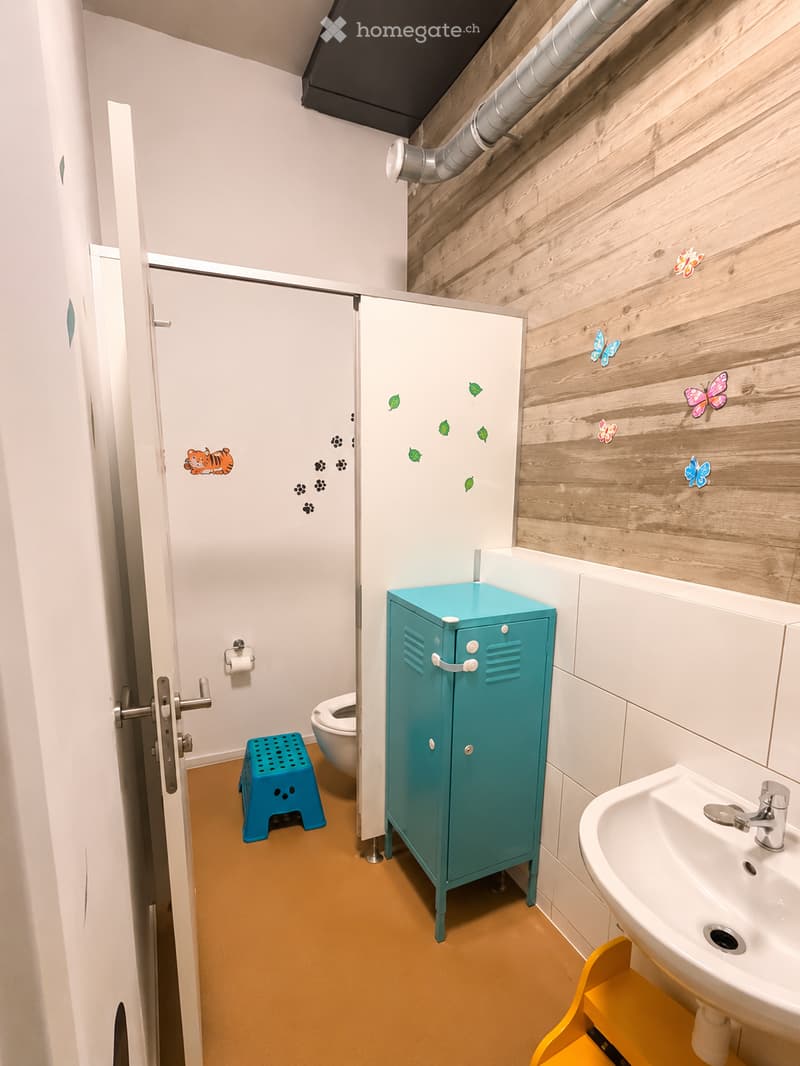 Bright, modern bathroom with a white sink, teal locker, blue and yellow stools, and a toilet. The walls feature playful decals including butterflies and leaves.