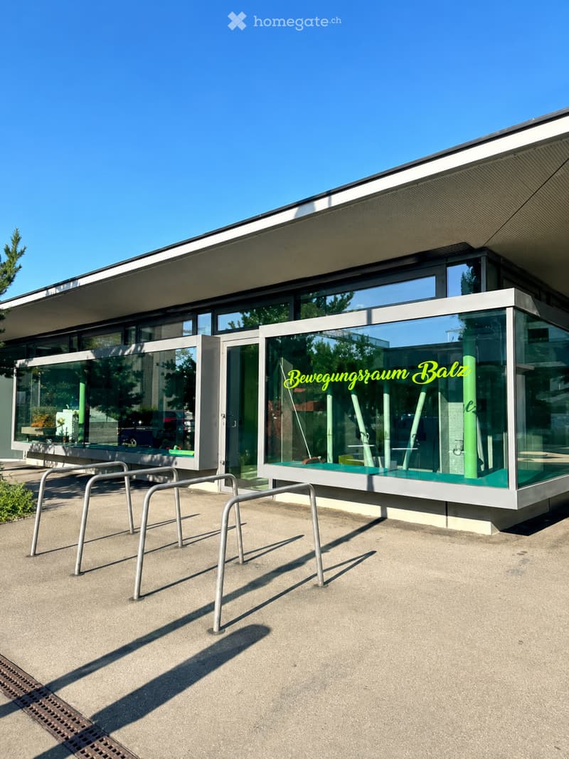 Modern building with large glass windows and a sleek, contemporary design. The entrance features a covered area with a bench and metal chairs.