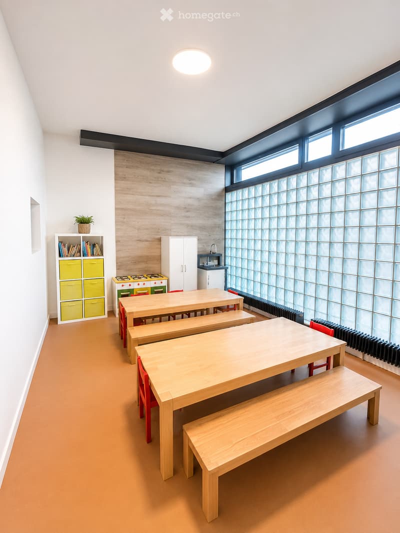 Brightly lit room with wooden tables and benches, red chairs, a small kitchen area with a sink and faucet, a white cabinet, a colorful shelf unit, and a small plant on top.