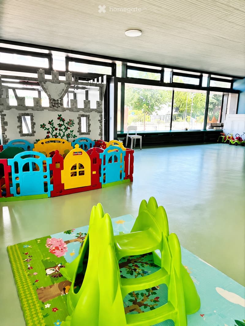 Brightly colored children's play area with a toy castle, toy fence, toy chairs, and a toy table. The room features large windows and a green play mat with a tree design.