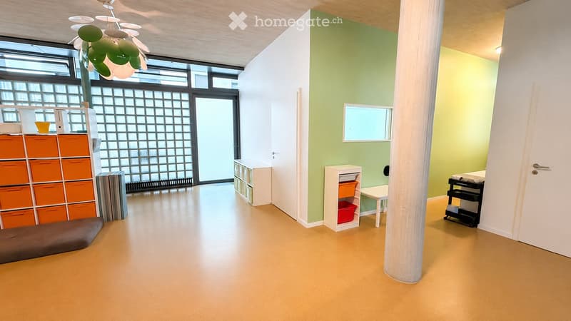 Bright, modern room with light green and white walls, wooden floor, and large windows. Features include a storage unit with orange drawers, a small table with a chair, and a cabinet with colorful bins.