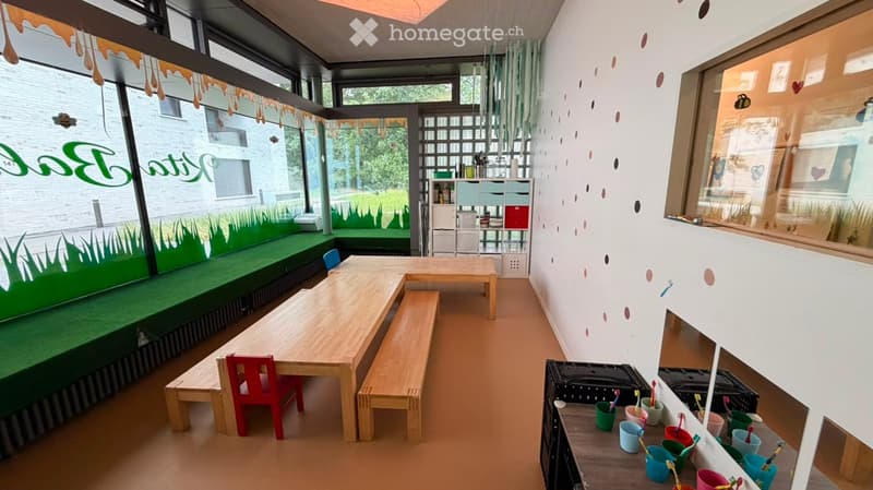Bright, playful room with large windows, green painted seating area, wooden tables and benches, colorful chairs, shelves with storage, and children's toys.