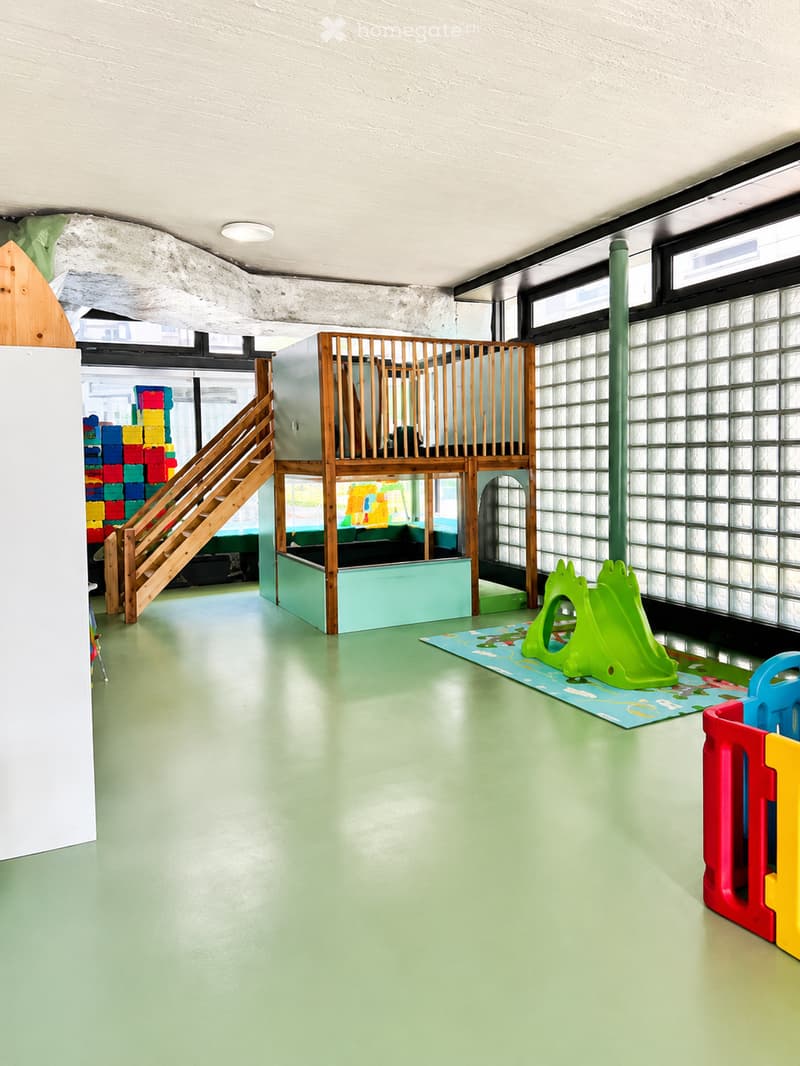 Brightly colored children's play area with a wooden staircase leading to an elevated platform, a play slide, colorful toy blocks, and a play mat with a toy chair.