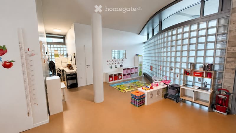 Bright, open room with large windows, wooden floor, and various children's furniture and toys including a desk, shelves, sink, and play area.