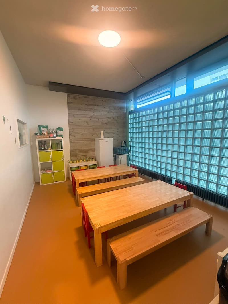 Bright room with wooden tables and benches, a refrigerator, a green shelf, and a large window with frosted glass panels.