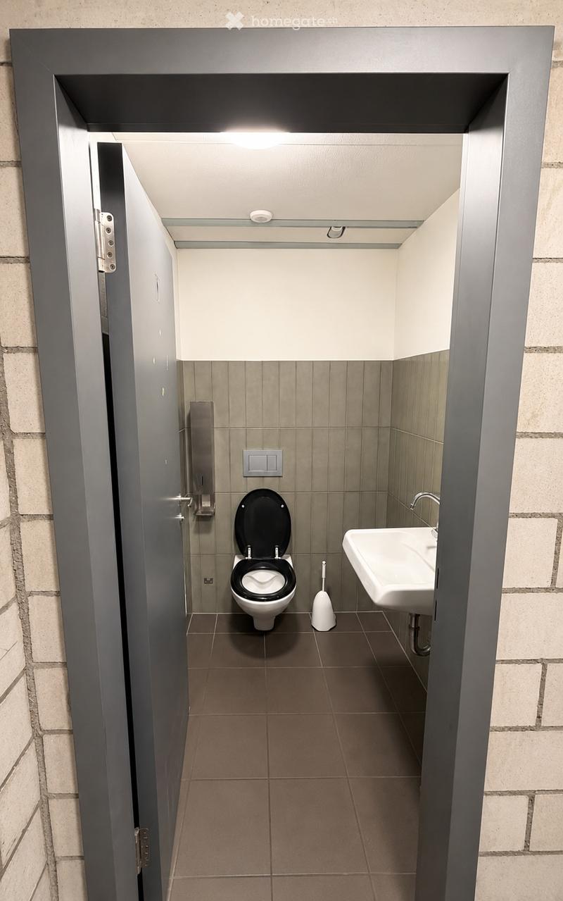 Modern bathroom with a toilet, sink, and toilet brush. The walls are tiled in a neutral color, and the floor is covered with brown tiles. The door is partially open, revealing the interior.