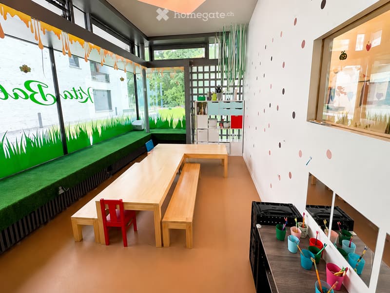 Brightly colored children's room with a large window, green carpeted seating area, wooden tables, benches, and colorful cups and pencils on a wooden shelf.