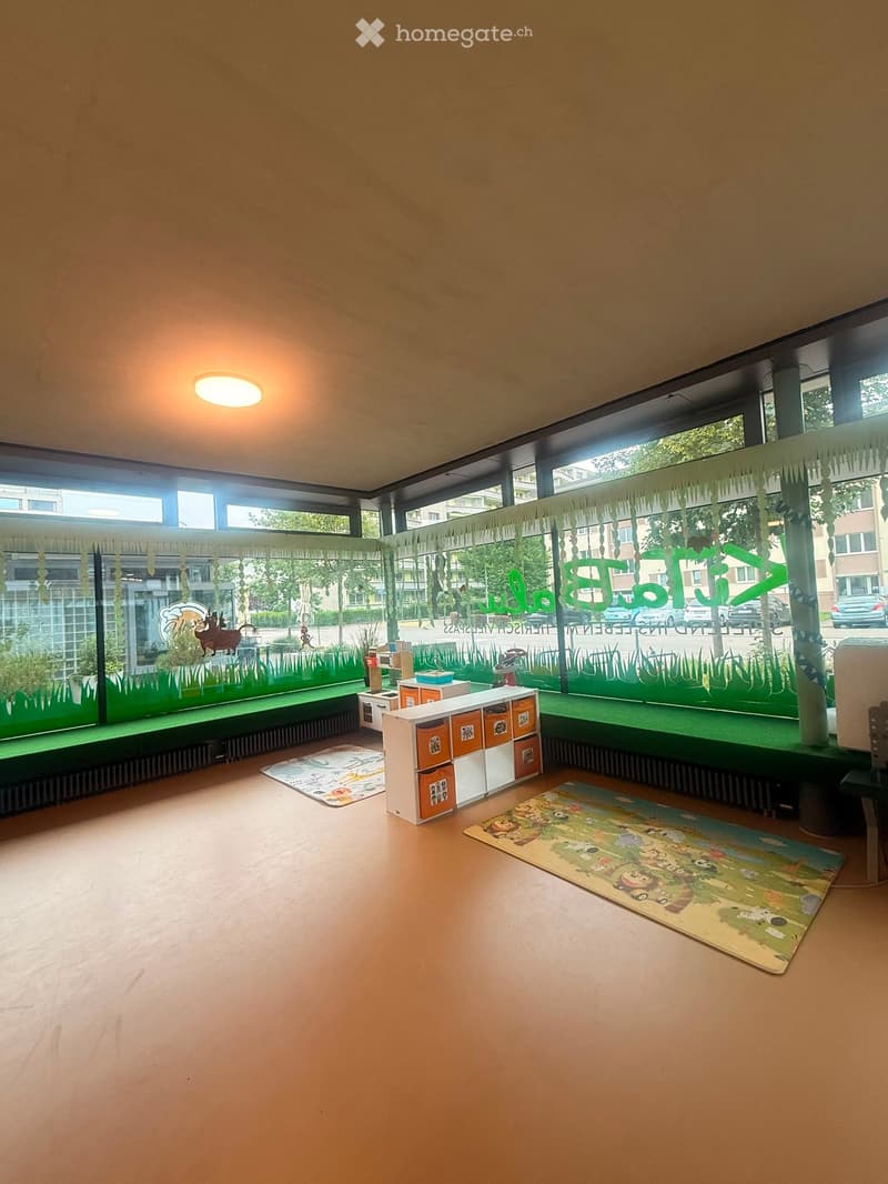 Brightly lit room with large windows, green play area, storage unit, colorful play mats, and a small table.