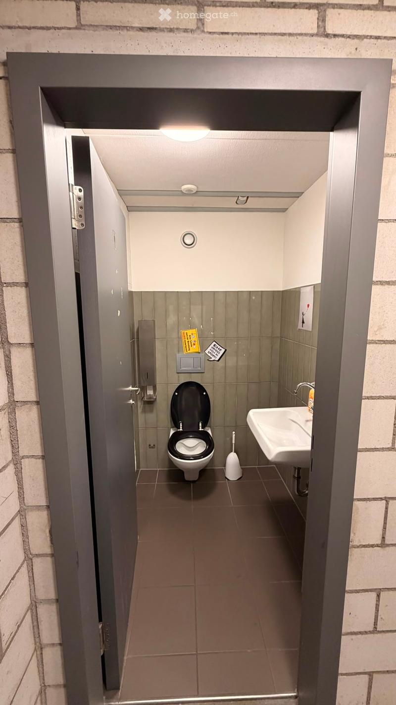 Compact bathroom with a toilet, sink, and toilet brush visible. The space features tiled walls and a dark tiled floor.