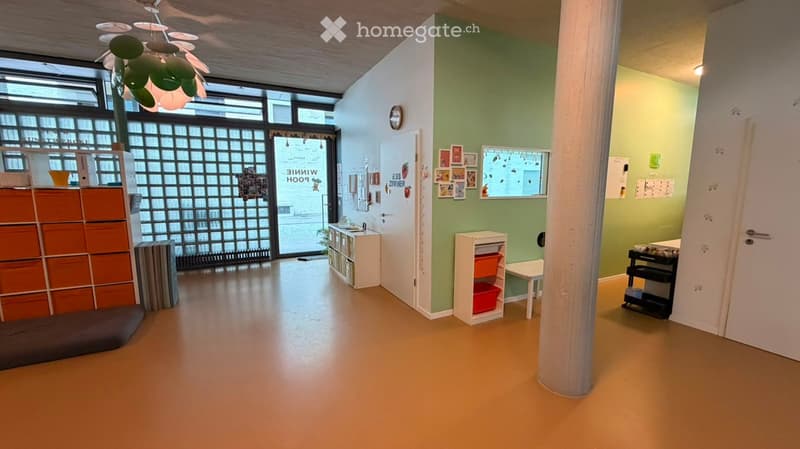 Bright, open room with light green walls, wooden floor, large windows, and a central column. Features include a storage unit, small table, chair, shelves, and a cabinet.