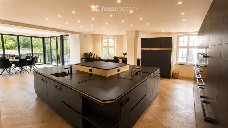 Modern kitchen with dark countertops, island, sink, and cabinets. Large windows and a balcony are visible.