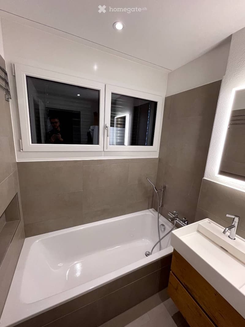 Modern bathroom with a white bathtub, a sink with a chrome faucet, and large windows with white frames. The walls are tiled in a combination of light and dark brown tiles.