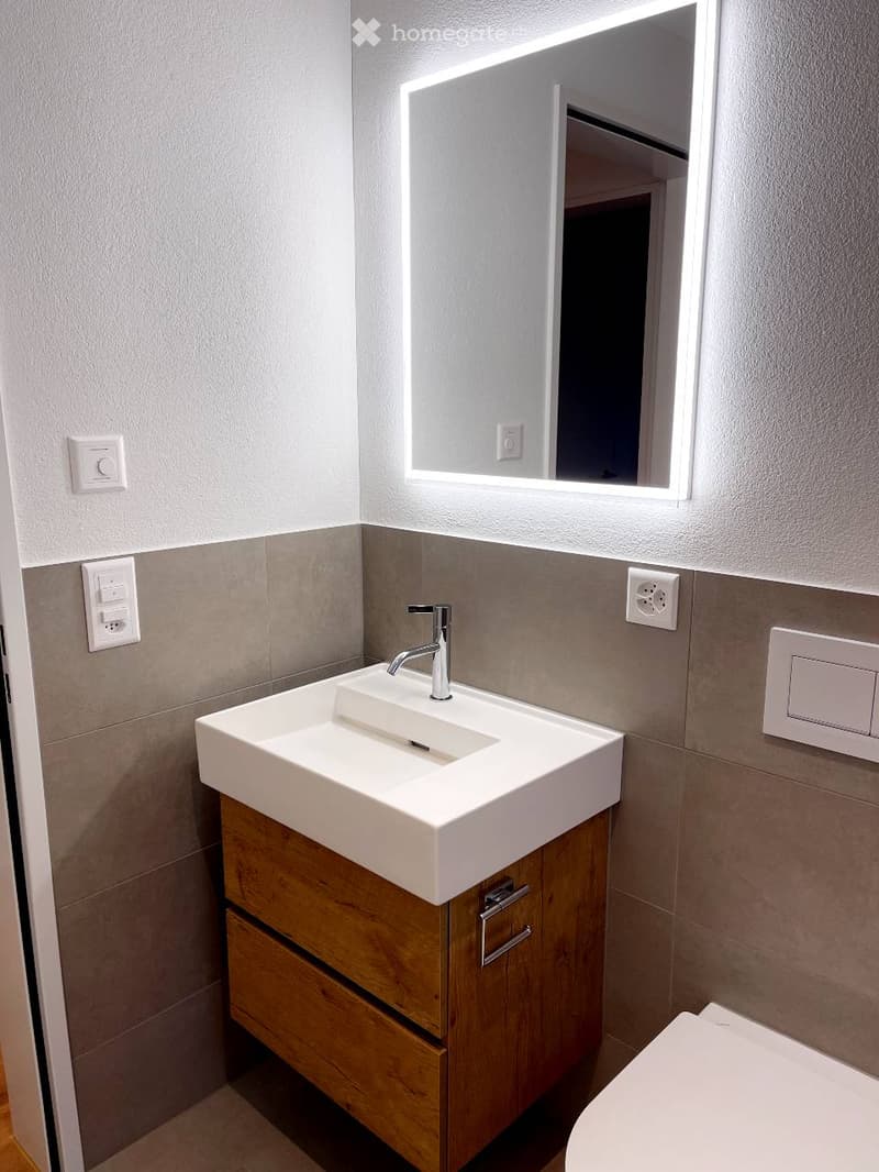Modern bathroom with a white sink, wooden cabinet, and illuminated mirror above it. The walls are tiled in a neutral color scheme, and the faucet is sleek and contemporary.