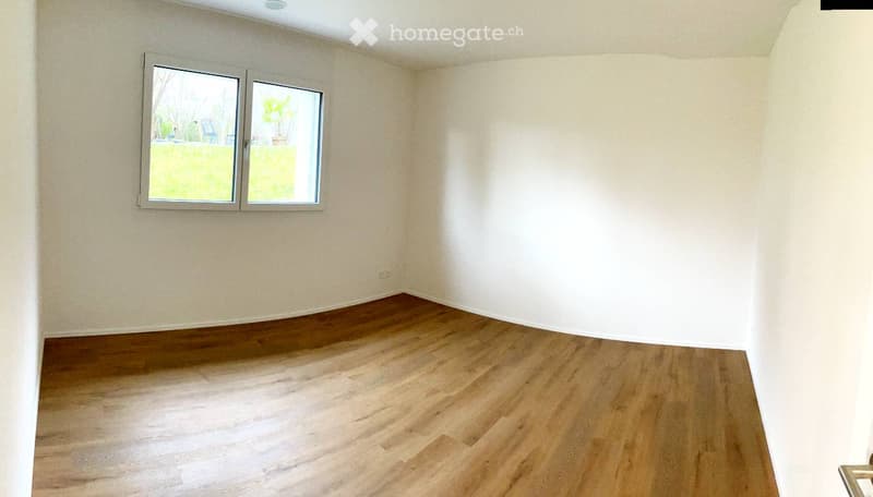 Empty room with light-colored walls and wooden flooring, featuring two windows with white frames and a view of a green outdoor area.