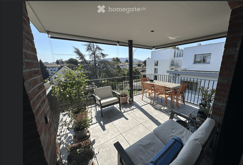 Balcony with a sofa, chairs, a table, and several potted plants, overlooking a cityscape with buildings and trees.